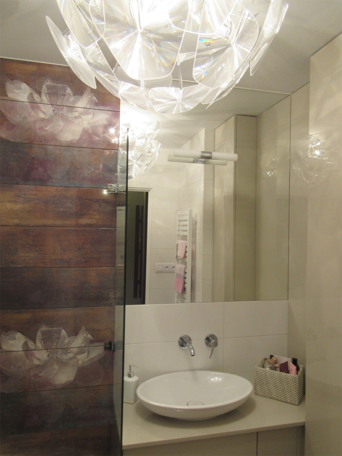 Nowoczesna łazienka z drewnopodobnymi płytkami z motywem kwiatowym na ścianie kabiny prysznicowej, owalną umywalką nablatową, chromowanymi bateriami i lustrem odbijającym grzejnik drabinkowy...