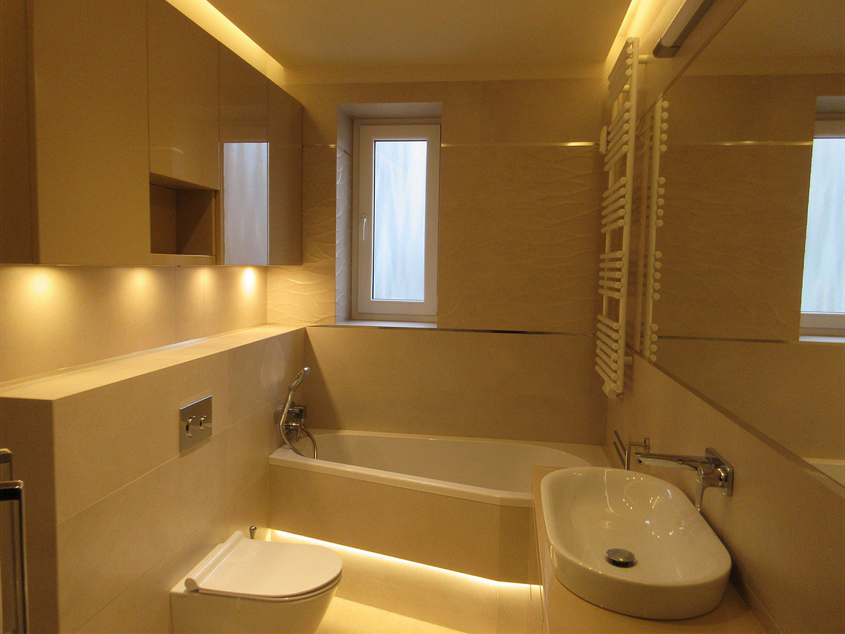 Nowoczesna łazienka w beżowych odcieniach z podświetleniem LED, wisząca toaleta, wanna z baterią, umywalka nablatowa i grzejnik drabinkowy na tle lustra.