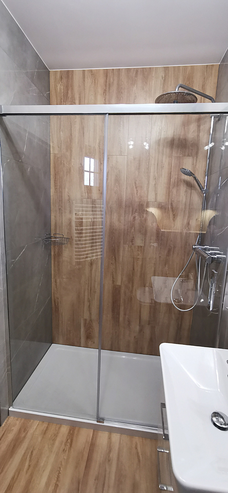 Kabina prysznicowa z przesuwnymi drzwiami, wyłożona płytkami imitującymi drewno i szarym marmurem, z widoczną armaturą i umywalką.