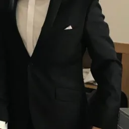 Uśmiechnięty mężczyzna w czarnym smokingu z białą koszulą i czarną muchą, stojący na tle ściany.