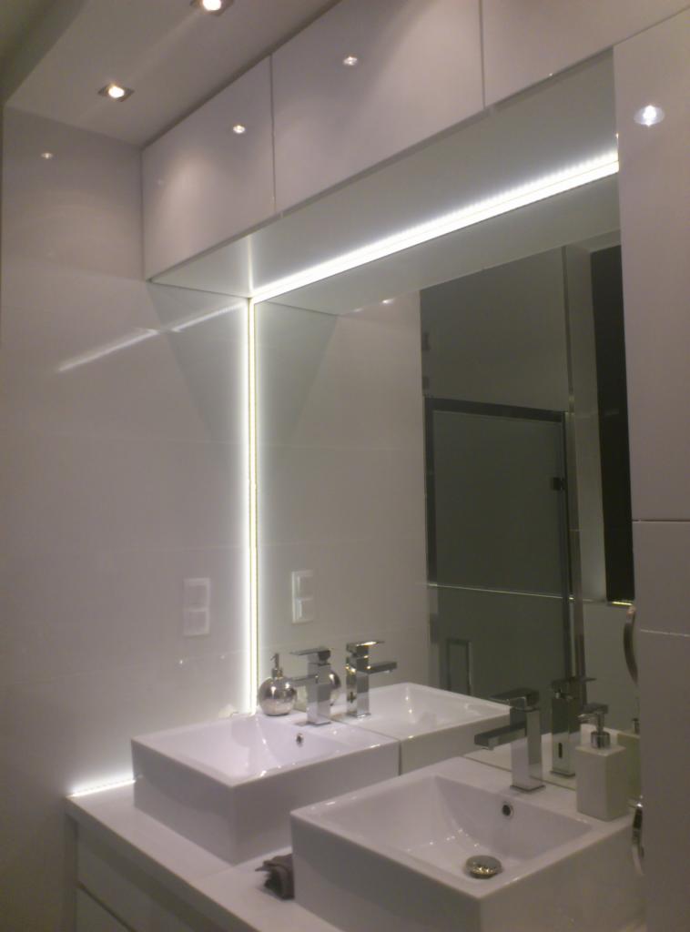 Nowoczesna łazienka z podwójną umywalką, białymi płytkami i oświetleniem LED. Minimalistyczny design i lustro z wbudowanym światłem. Widoczne detale armatury.