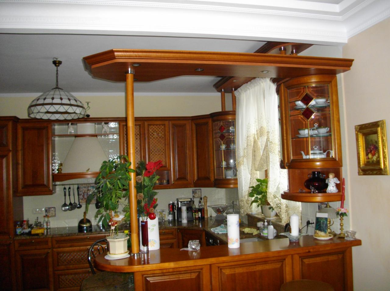 Kuchnia z drewnianą zabudową i barkiem, oświetlona lampą witrażową. Dekoracje roślinne i zasłony dodają przytulności. Widoczne szklane witryny i akcesoria kuchenne.