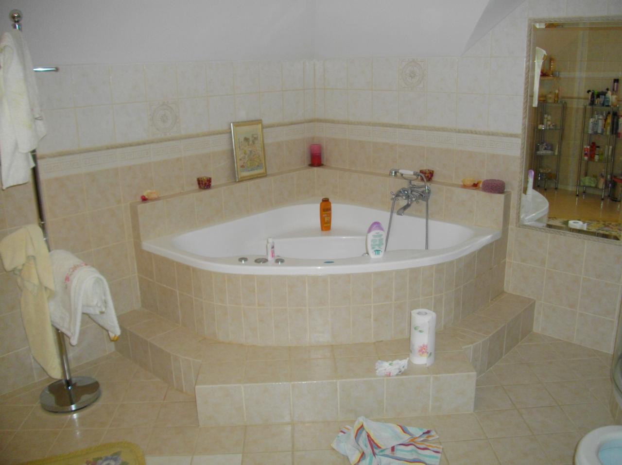 Jasna łazienka z narożną wanną obudowaną beżowymi płytkami, lustro z widokiem na półki z kosmetykami i ręcznik na stojaku. Ukończona aranżacja.