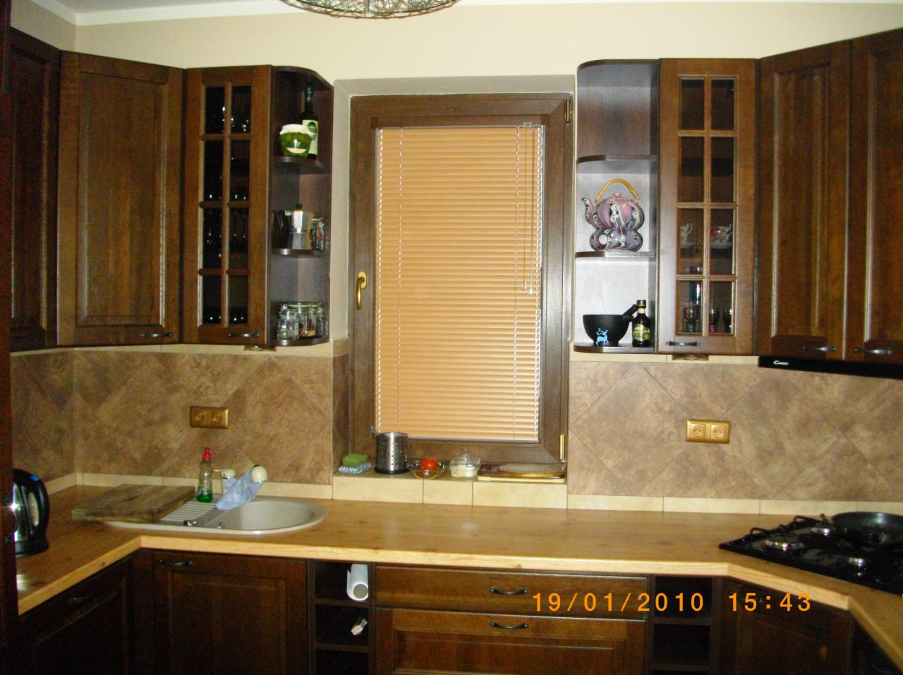 Kuchenne szafki z ciemnego drewna, brązowe płytki na ścianie, okno z żaluzjami i drewnianą ramą. Widok na blat z umywalką i kuchenką. Ciepłe oświetlenie.