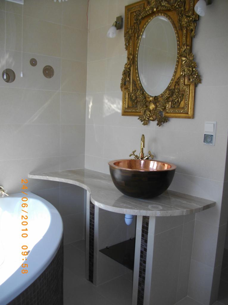 Elegancka łazienka z umywalką w kształcie misy, złotą armaturą i ozdobnym lustrem w złotej ramie. Blat z marmuru, płytki w odcieniach beżu. Luksusowe wykończenie.