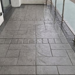 Zakład Remontowo-Budowlany - Balkon z ciemnoszarą posadzką z betonu stemplowanego, imitującą kamienne płyty, z mokrą powierzchnią, obok białej ściany i nowoczesnej balustrady ze szkła i stali.