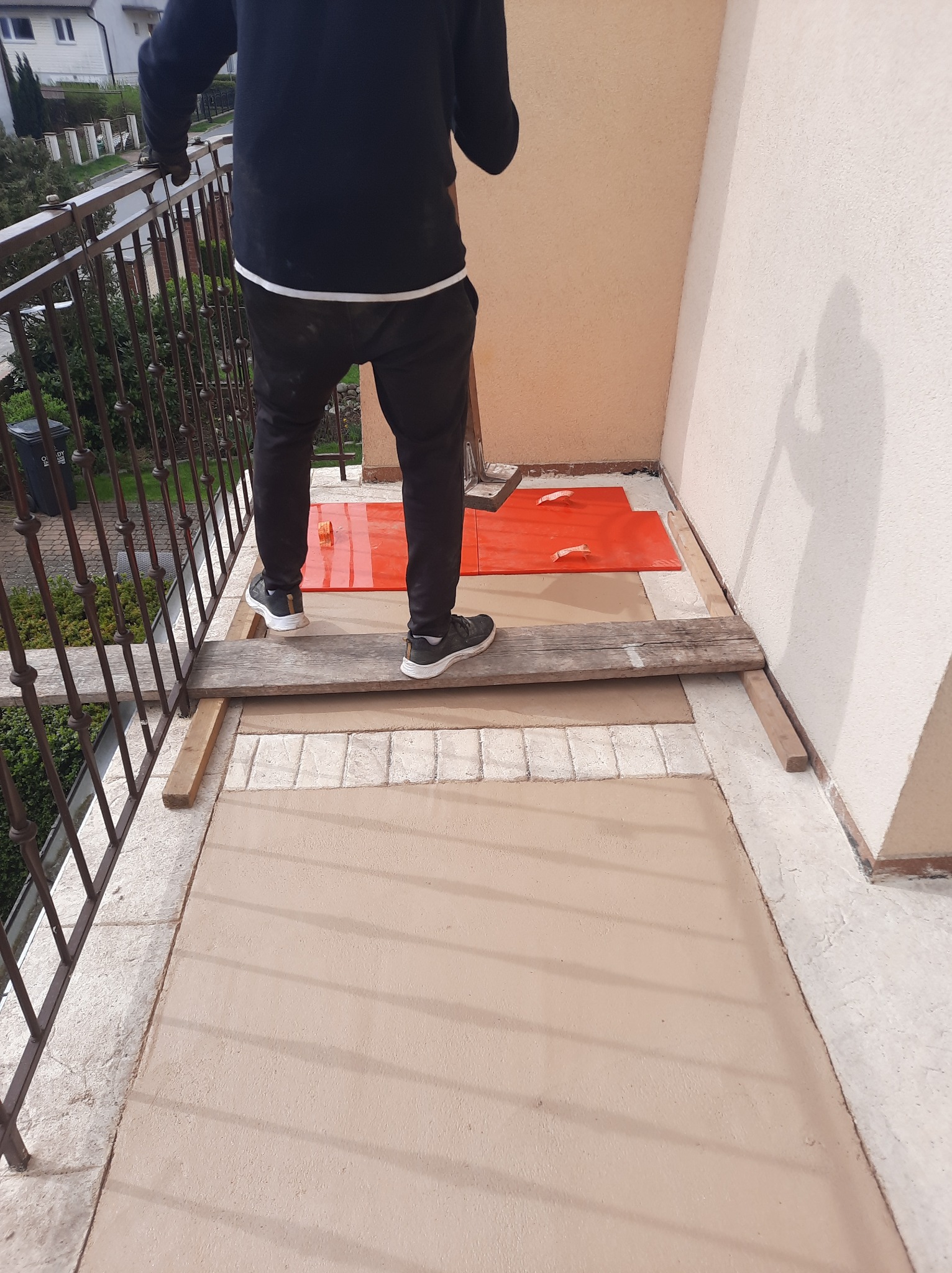 Balkon w trakcie prac: świeżo wylana, beżowa posadzka, zabezpieczona część podłoża. Widoczne narzędzia i fragment osoby wykonującej prace.