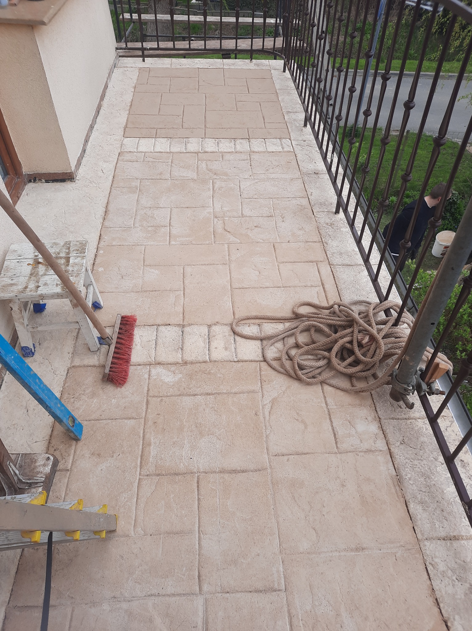 Balkon z nową, beżową posadzką imitującą kamień. Widoczne narzędzia: drabina, szczotka, sznurek. Metalowa balustrada i fragment elewacji w tle.