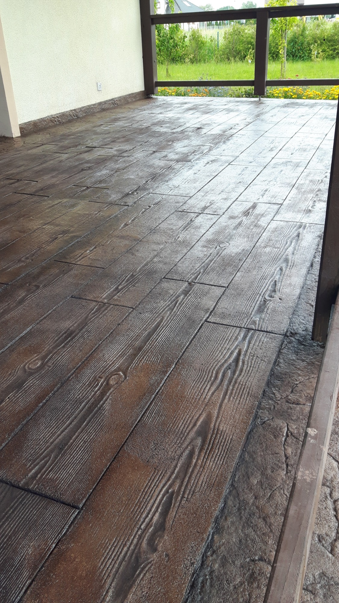 Balkon z podłogą imitującą drewniane deski, wykonaną z szarego betonu z widoczną fakturą słojów drewna i nieregularną krawędzią. Balustrada w tle i zieleń za nią.