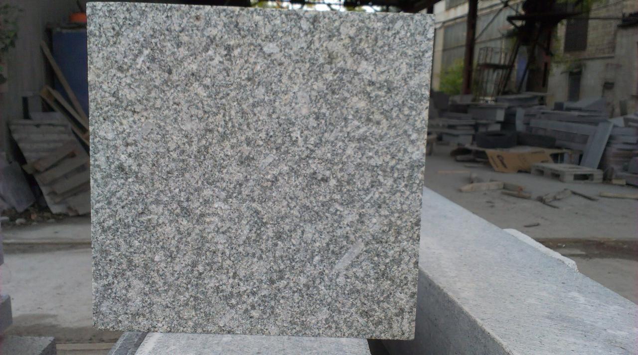Szara, kwadratowa płyta granitowa o ziarnistej fakturze, ułożona na krawędzi, z widocznymi elementami składowiska w tle.