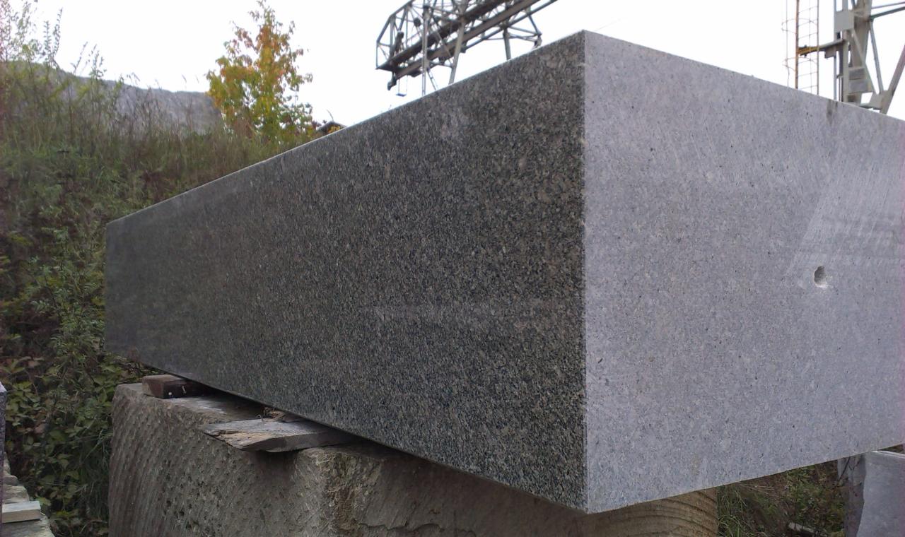 Szary, prostokątny blok kamienny z widoczną fakturą, ustawiony na mniejszym bloku. W tle zieleń i konstrukcja stalowa.