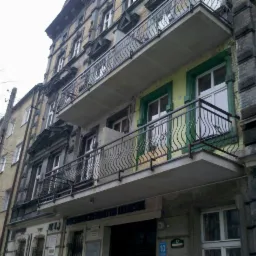 Budynek mieszkalny z balkonami zabezpieczonymi metalowymi balustradami o falistym wzorze, widoczne detale architektoniczne i zróżnicowana kolorystyka elewacji.