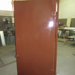 Brązowe, stalowe drzwi warsztatowe, świeżo pomalowane proszkowo, widoczne zawiasy i zamek, ustawione na podkładkach w hali produkcyjnej.