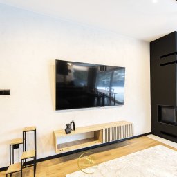 MYSprojekt Marek Myszkowski - Jasne wnętrze z telewizorem, drewnianą szafką RTV i czarnym kominkiem z dekoracją czaszki. Minimalistyczny design z drewnianą podłogą i jasnym dywanem.