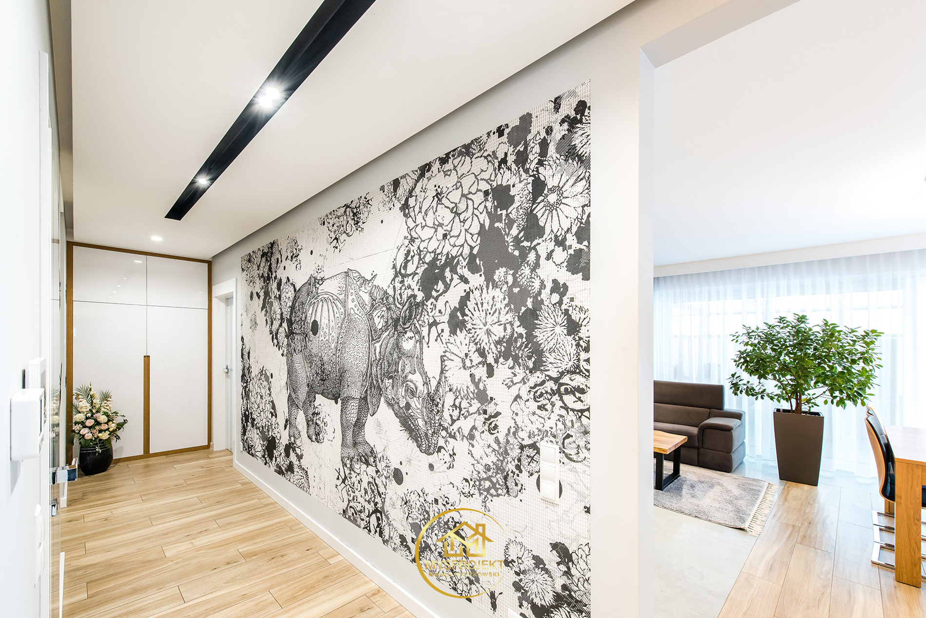 Nowoczesny przedpokój z fototapetą w motywy roślinne i zwierzęce, drewnianą podłogą i białymi szafami w zabudowie. Widok na salon z rośliną doniczkową.