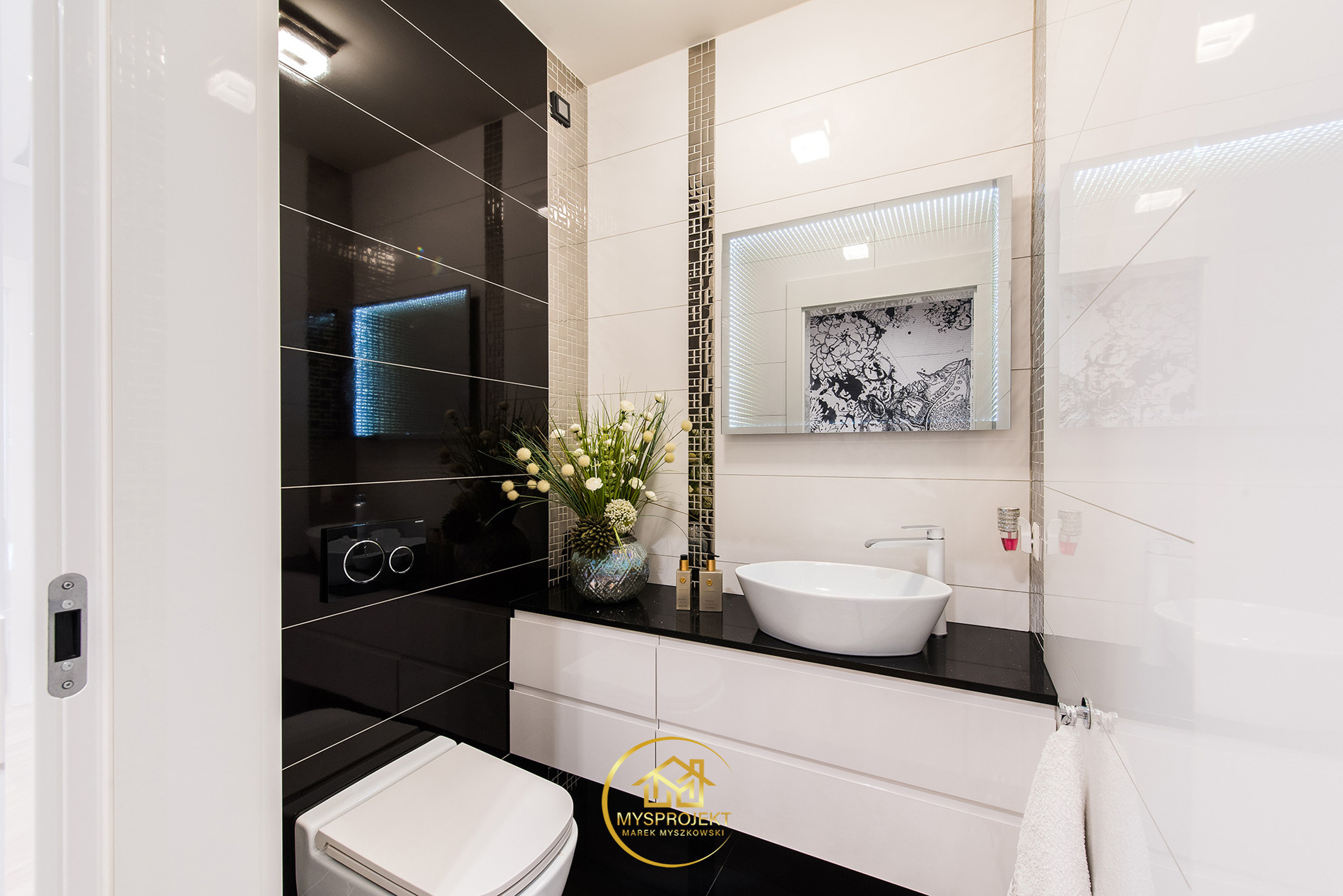 Nowoczesna łazienka z czarno-białymi płytkami, podświetlanym lustrem i umywalką nablatową. Kwiaty w wazonie dodają elegancji. Widoczny fragment toalety.