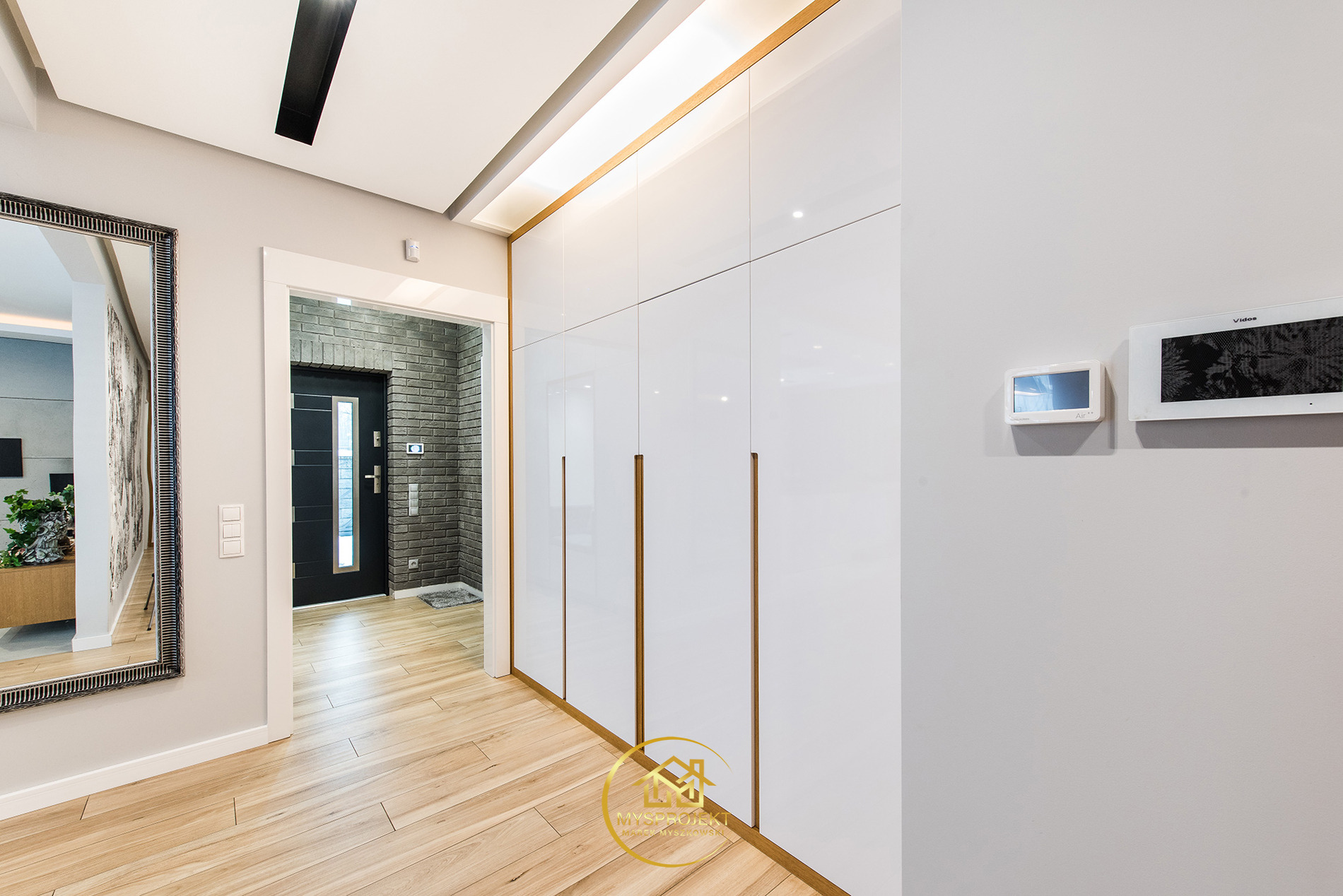 Nowoczesny przedpokój z białymi szafami w zabudowie, drewnianą podłogą i dużym lustrem. Widoczny fragment wejścia z drzwiami i ceglaną ścianą.
