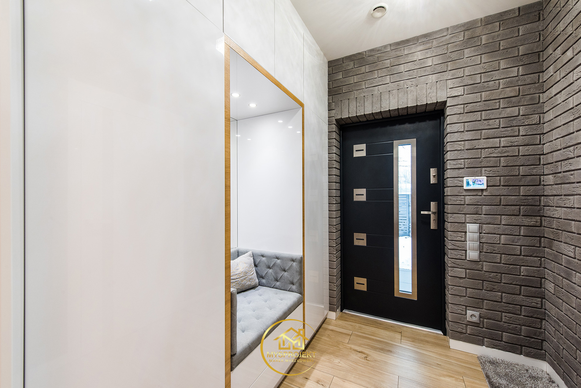 Nowoczesny przedpokój z siedziskiem w zabudowie, ceglana ściana i czarne drzwi wejściowe. Jasne drewno na podłodze. Minimalistyczny design.