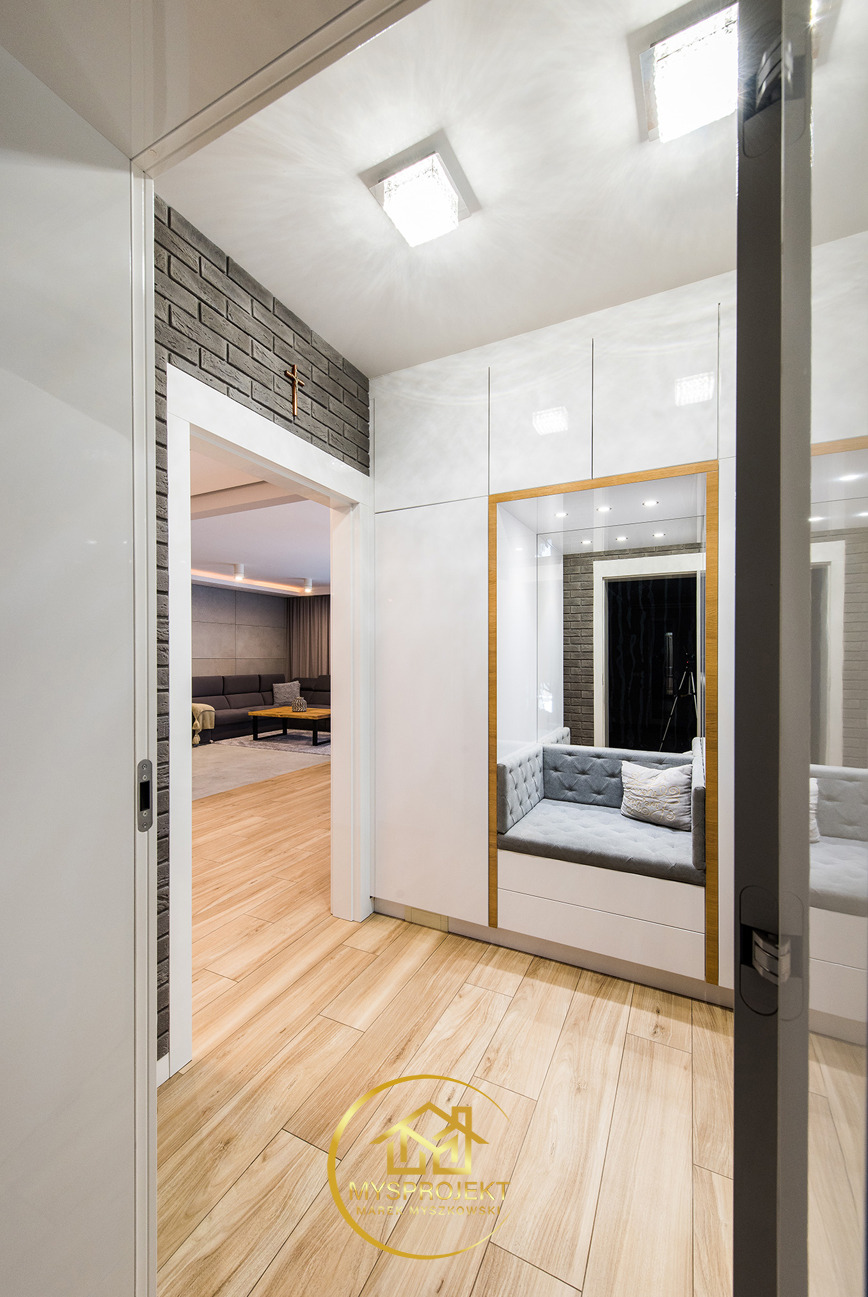 Nowoczesny przedpokój z siedziskiem w zabudowie, cegłą dekoracyjną i drewnianą podłogą. Widok na salon przez otwarte drzwi. Lustro odbija wnętrze.