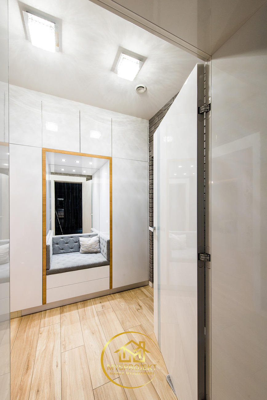 Biała zabudowa meblowa z siedziskiem wnękowym z szarą tapicerką i poduszką, drewniana podłoga, oświetlenie sufitowe. Nowoczesny design wnętrza.