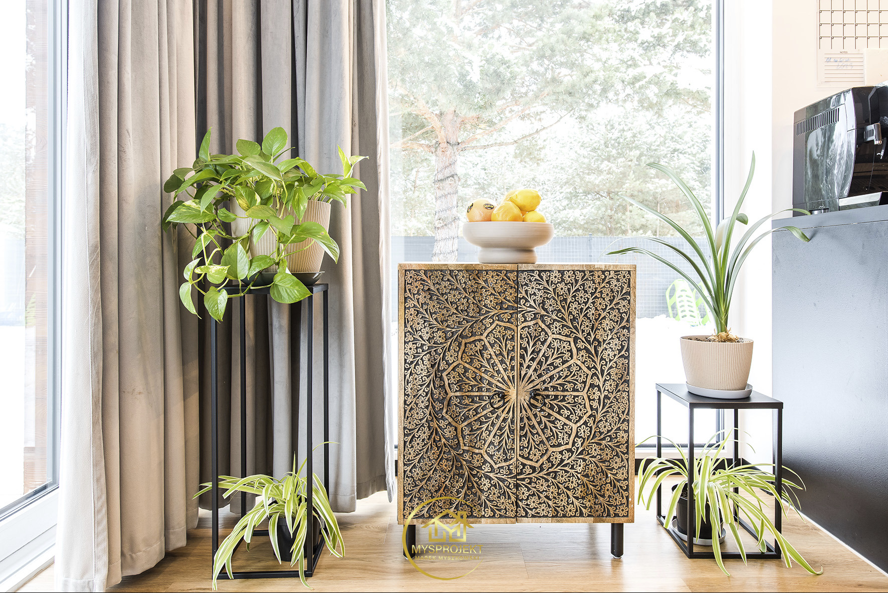 Stylowa komoda z rzeźbionym wzorem kwiatowym, zwieńczona misą z cytrynami. Obok rośliny doniczkowe na metalowych stojakach, okno z widokiem na drzewa.
