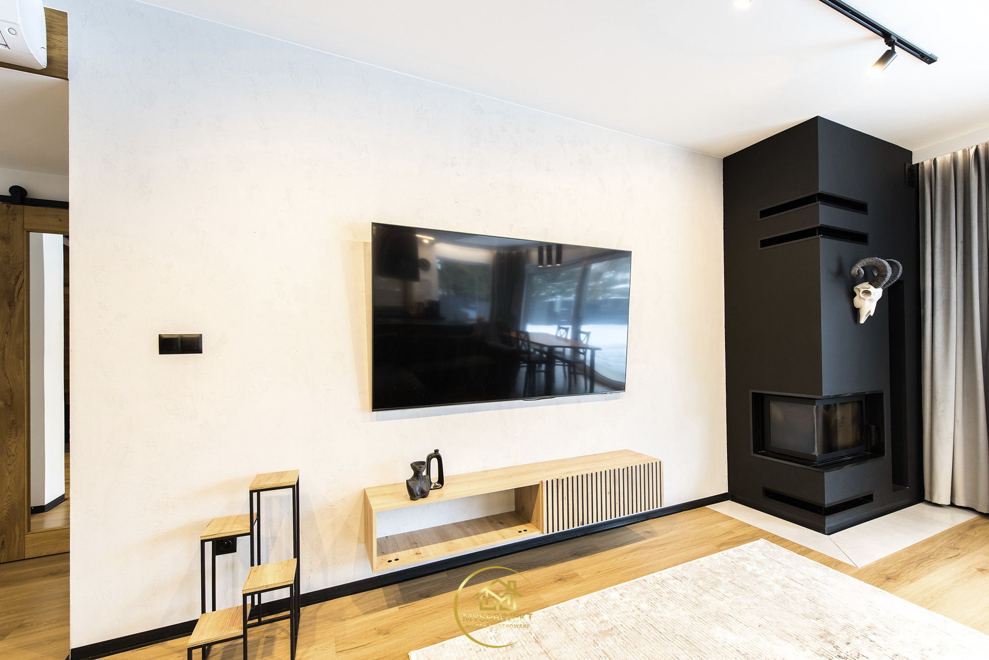 Jasne wnętrze z telewizorem, drewnianą szafką RTV i czarnym kominkiem z dekoracją czaszki. Minimalistyczny design z drewnianą podłogą i jasnym dywanem.