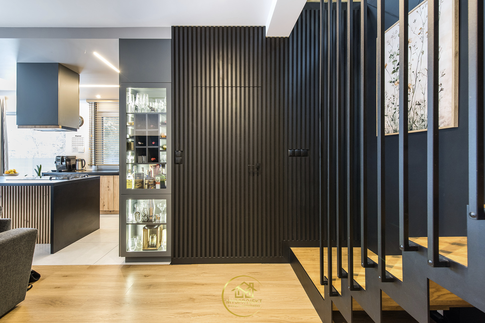 Nowoczesne wnętrze: czarna ściana z lamelami, przeszklona witryna z alkoholem i szkłem, drewniane schody z czarną balustradą, parkiet. Widok na kuchnię.