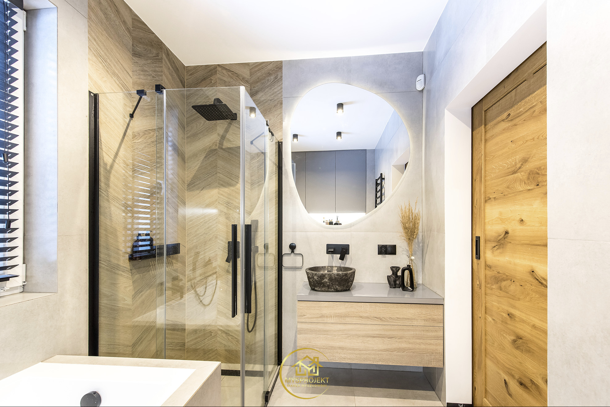 Nowoczesna łazienka z kabiną prysznicową typu walk-in, umywalką z kamienia i owalnym lustrem z podświetleniem LED. Drewniane akcenty i neutralna kolorystyka.