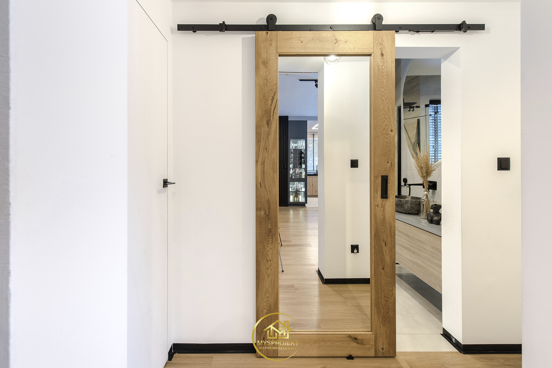 Drewniane drzwi przesuwne w stylu rustykalnym, zamontowane na czarnej szynie, oddzielają dwa pomieszczenia. Widok na łazienkę i salon. Minimalistyczny design.