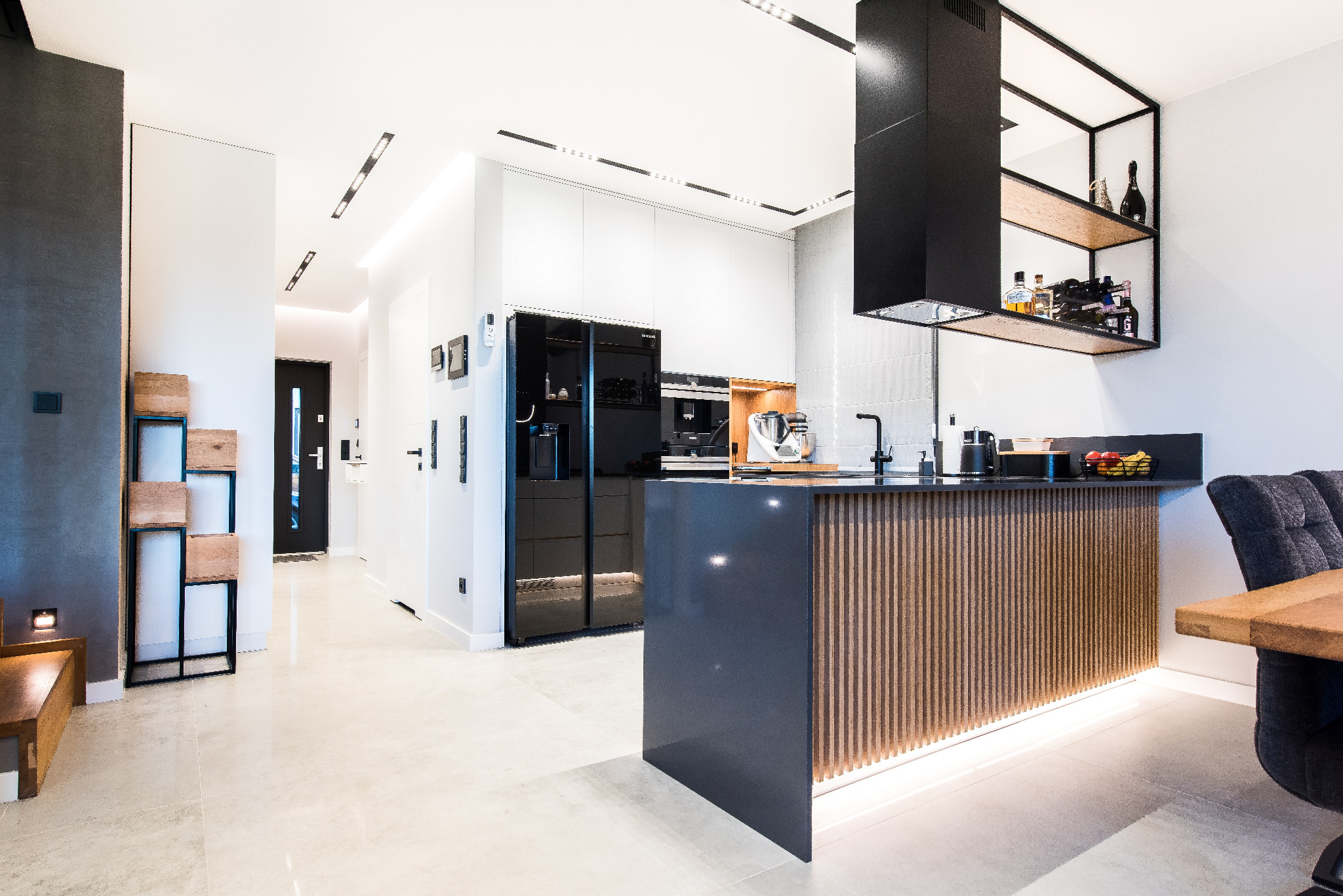 Nowoczesna kuchnia z wyspą, czarnymi akcentami i oświetleniem LED. Minimalistyczny design, drewniane elementy dekoracyjne, sprzęt AGD w zabudowie. Widok na korytarz.