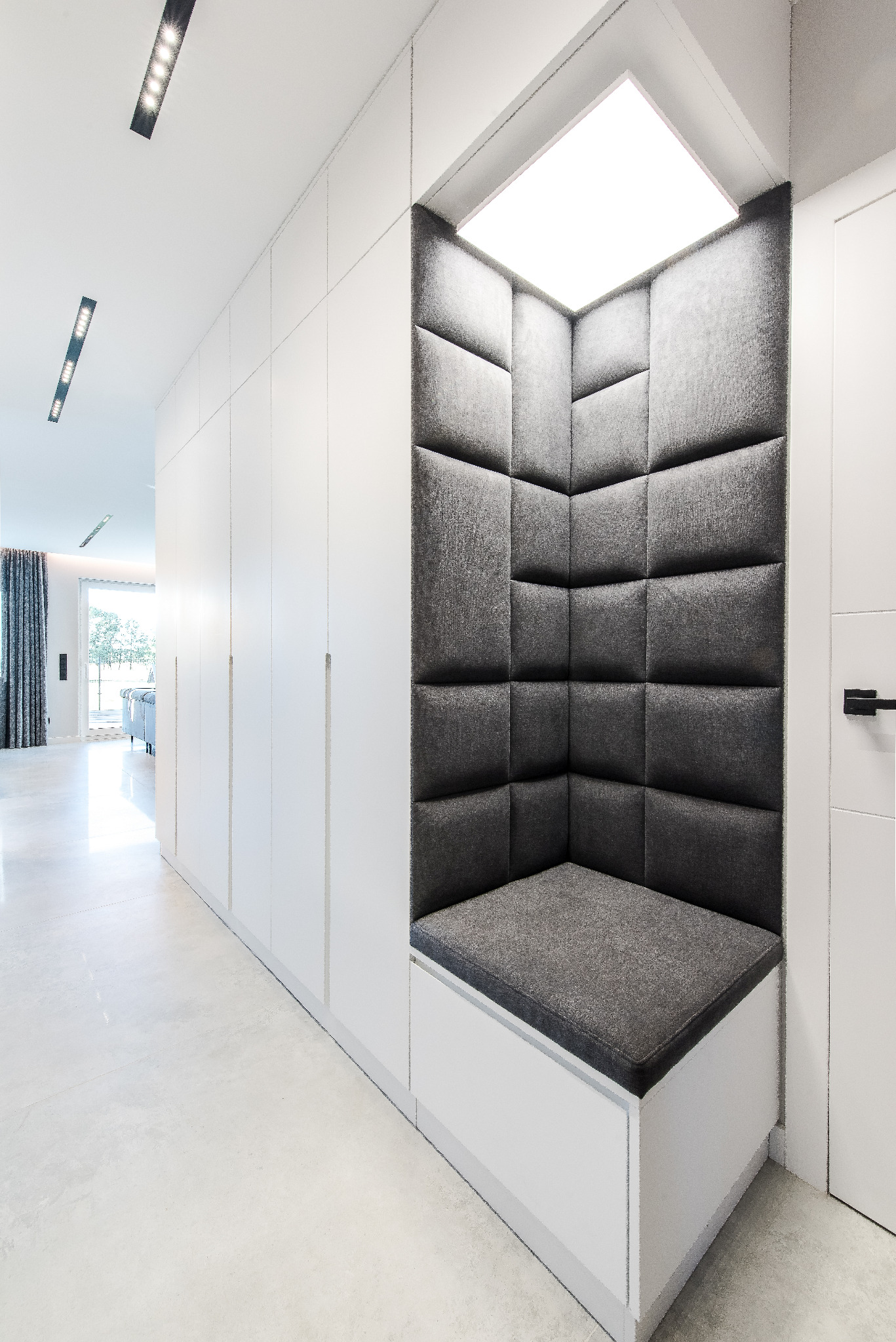 Nowoczesny przedpokój z białą zabudową szaf i wnęką do siedzenia wyłożoną szarą tapicerką. Minimalistyczne oświetlenie sufitowe.