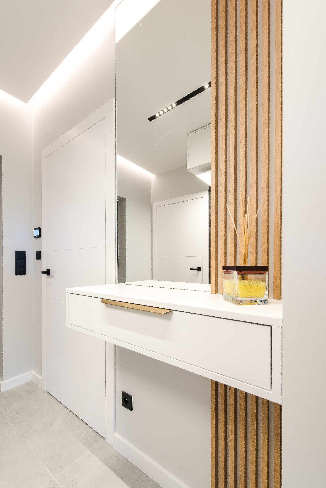Nowoczesny przedpokój z białymi meblami, lustrem i drewnianymi lamelami. Na blacie dekoracyjny dyfuzor zapachowy. Minimalistyczny design, jasne wnętrze.