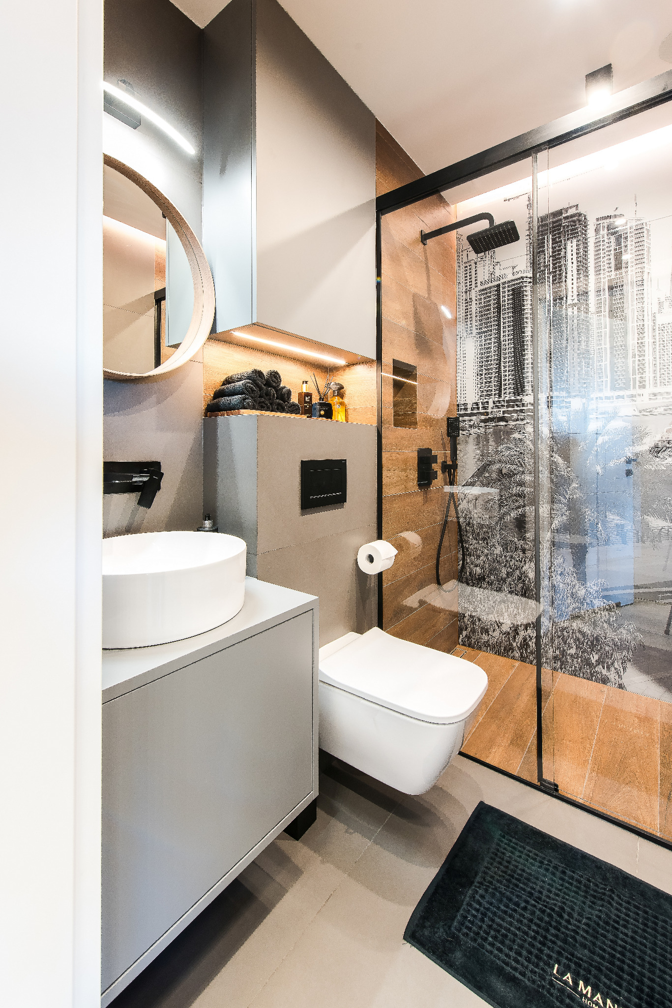 Nowoczesna łazienka z okrągłym lustrem, umywalką nablatową, podwieszaną toaletą i kabiną prysznicową z motywem miejskim. Stonowana kolorystyka, oświetlenie LED.