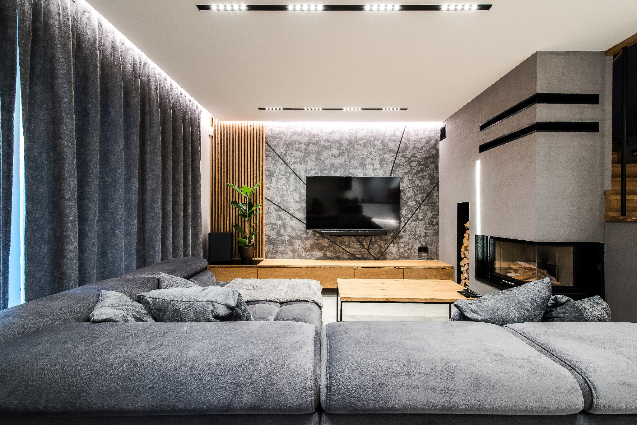 Nowoczesny salon z szarą sofą, telewizorem na ścianie o betonowej fakturze i kominkiem. Drewniane lamele i oświetlenie LED dodają ciepła wnętrzu.