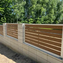 Nowoczesne ogrodzenie z poziomych paneli imitujących drewno, osadzone na betonowych słupkach i podmurówce z szarej kostki, z zielonym tłem drzew.