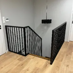 Nowoczesna, czarna balustrada schodowa z pionowymi prętami, na drewnianych schodach i parkiecie, oświetlona przez czarny, cylindryczny żyrandol w minimalistycznym wnętrzu.