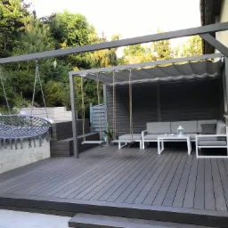 Zadaszony taras z szarej deski kompozytowej, wiszące huśtawki i meble ogrodowe w nowoczesnym stylu, widok na zieleń i fragment budynku.