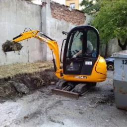 Żółta mini koparka JCB 8014 CTS w trakcie prac ziemnych, operator w kabinie, kontener na gruz w tle. Widoczne ślady opon i wykopany teren.
