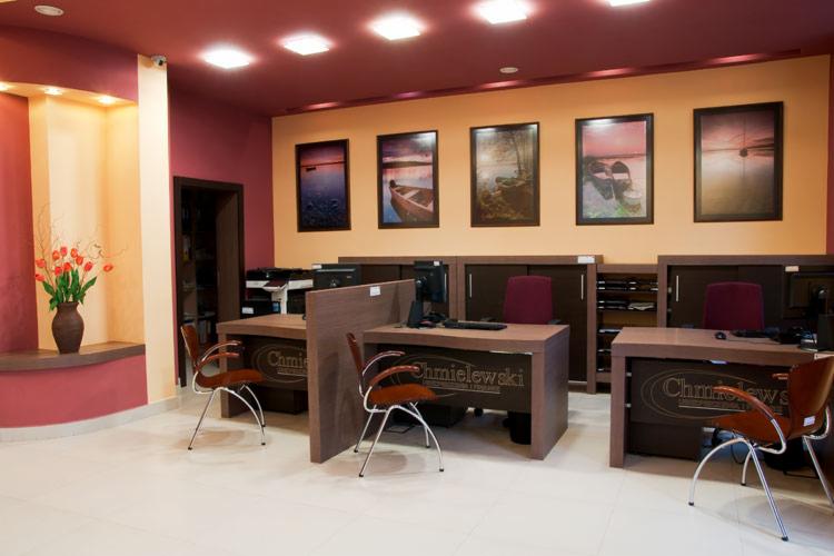 Wnętrze biura z trzema stanowiskami pracy, obrazami na ścianie i kwiatami w wazonie, utrzymane w ciepłej kolorystyce.