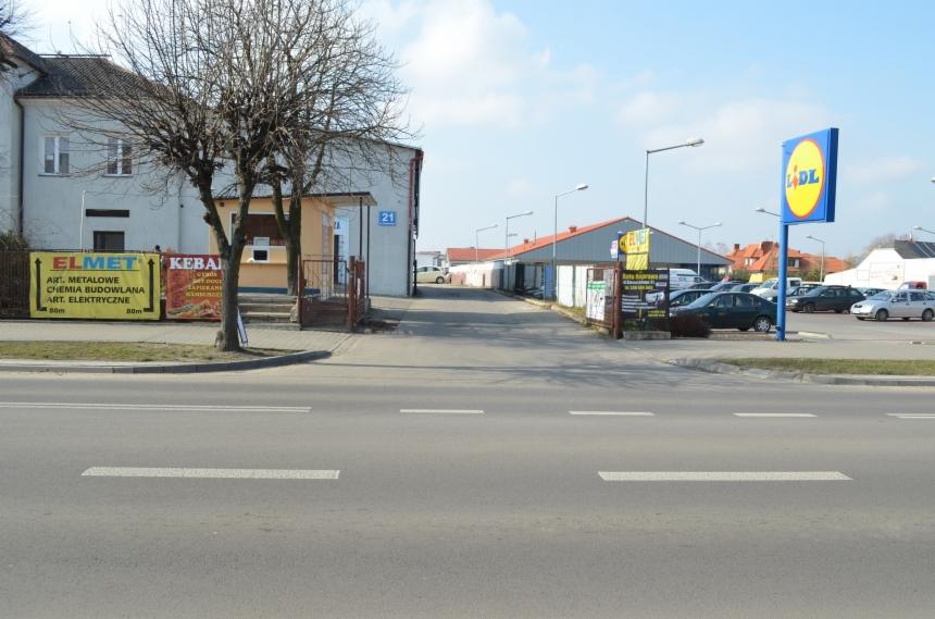 Ulica z budynkami, szyldami reklamowymi i parkingiem. W tle widoczny znak Lidl. Ujęcie z poziomu jezdni, w słoneczny dzień.