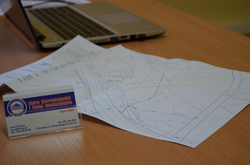 Plan geodezyjny rozłożony na biurku obok wizytówki firmy BNIUG i laptopa. Widoczne dane kontaktowe i adres firmy.