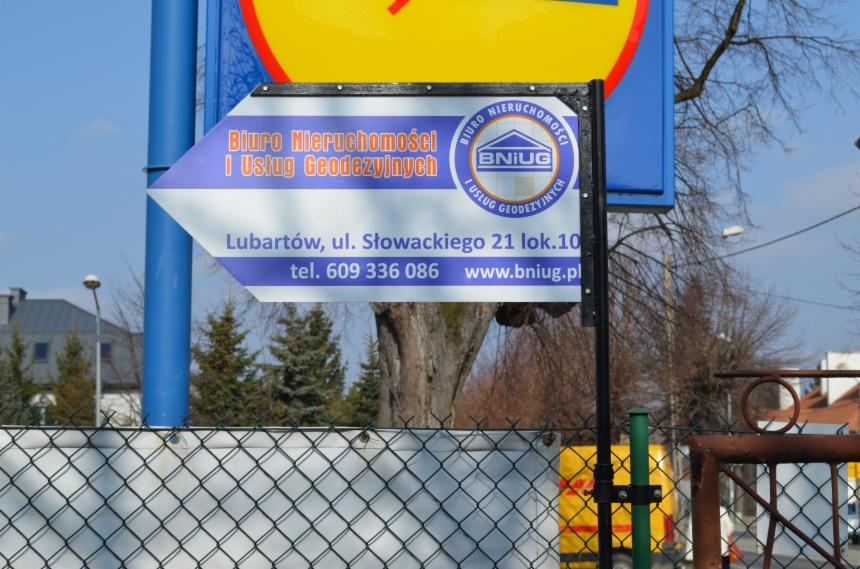 Tablica informacyjna Biura Nieruchomości i Usług Geodezyjnych BNIUG w Lubartowie, z adresem, numerem telefonu i adresem strony internetowej. Widok zza ogrodzenia.