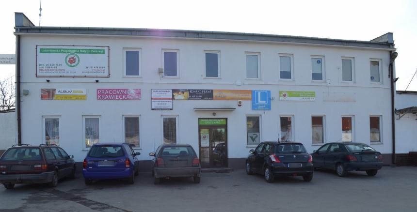 Biały, dwukondygnacyjny budynek z reklamami różnych firm i zaparkowanymi samochodami. Widoczne szyldy, okna i drzwi wejściowe.