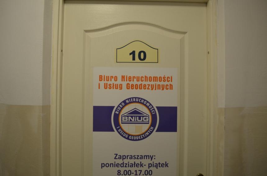 Drzwi z numerem 10 i informacją o Biurze Nieruchomości i Usług Geodezyjnych BNIUG. Na plakacie godziny otwarcia biura. Widok z bliska, oświetlenie sztuczne.