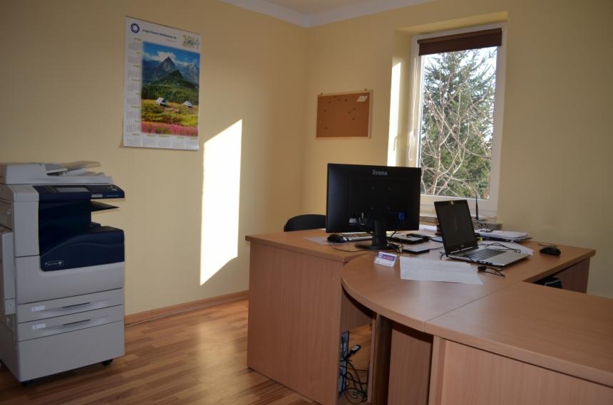 Biuro z biurkiem, komputerem, laptopem, drukarką i kalendarzem na ścianie. Widok na drzewa za oknem. Jasne ściany, drewniana podłoga.