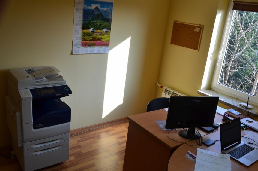 Wnętrze biura z biurkiem, komputerem, drukarką i kalendarzem ściennym z motywem górskim. Widok z okna na drzewa bez liści.