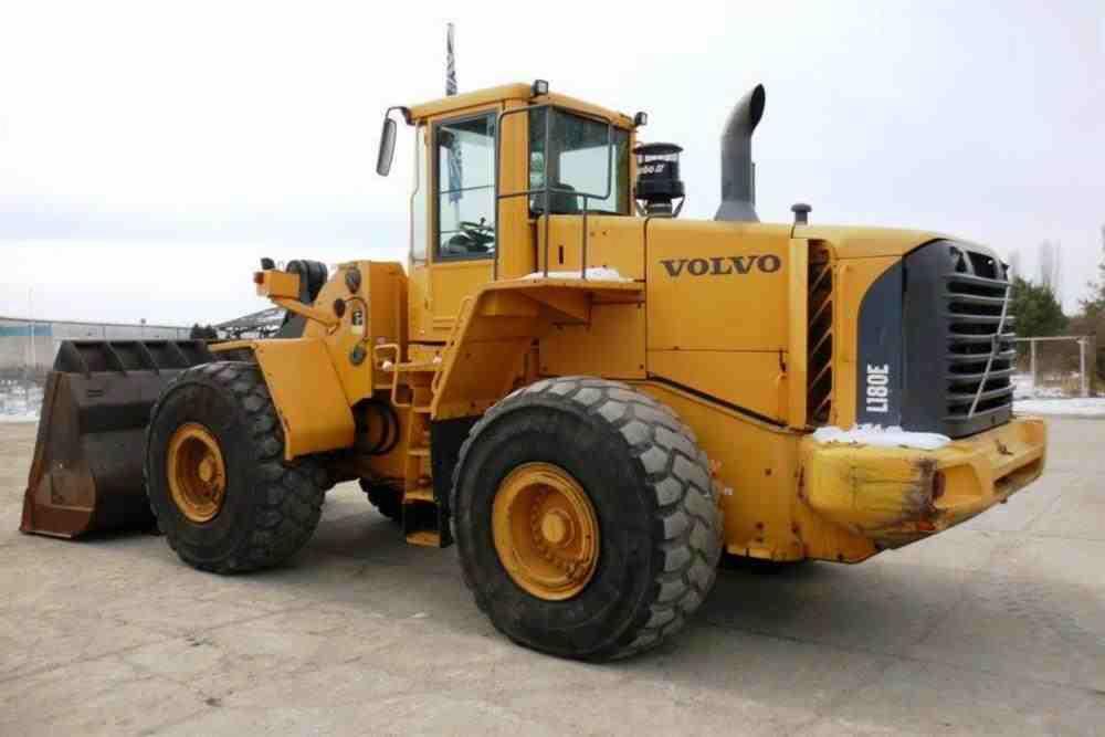 Żółta ładowarka kołowa Volvo L180E z widocznymi śladami użytkowania na przednim zderzaku, stojąca na betonowej powierzchni w pochmurny dzień.