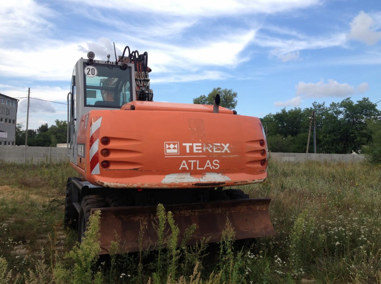 Pomarańczowa koparka kołowa Terex Atlas z widocznym numerem 20 na tle letniego krajobrazu z trawami i budynkiem w oddali.