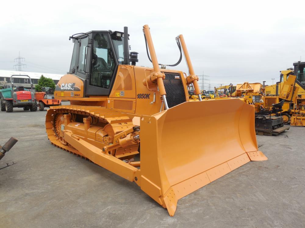 Pomarańczowa spycharka Case 1850K z szerokim lemieszem na placu budowy, widoczne inne maszyny w tle.