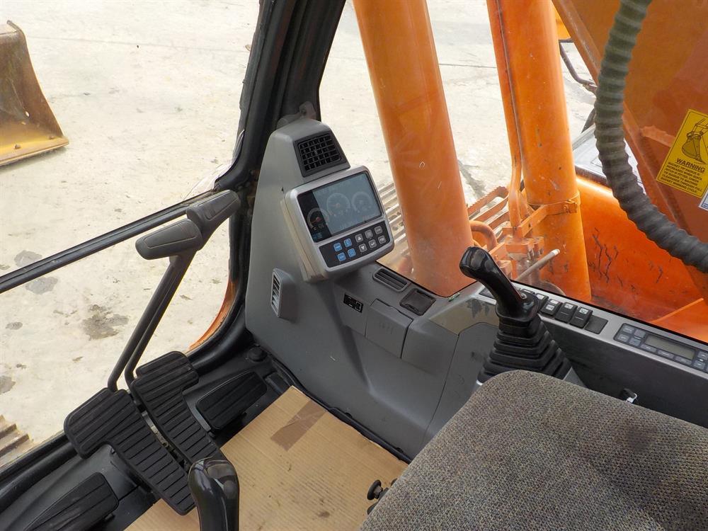 Wnętrze kabiny pomarańczowej koparki Doosan DX225LC z widocznymi pedałami, panelem sterowania i joystickiem.